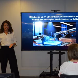 Palestrante apresenta o tema “O Código da Lei nº 14.634/2023: A Chave de Leitura das Normas de Contratações Administrativas na Bahia”. Ela está de pé ao lado de uma tela que exibe uma imagem ilustrativa com braços robóticos manuseando dados digitais, indicando abordagem tecnológica no contexto jurídico. O público acompanha sentado, atento à exposição.