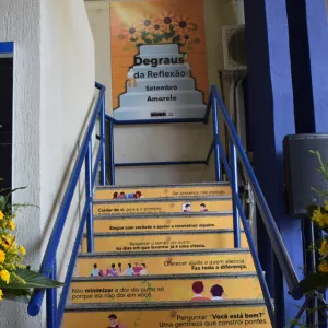 Escada decorada com frases de incentivo à empatia, escuta e cuidado emocional, como parte da campanha “Degraus da Reflexão – Setembro Amarelo”. Cada degrau traz uma mensagem positiva, estimulando atitudes simples que fazem a diferença no dia a dia. Ao fundo, há um painel com o nome da campanha.