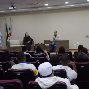 A imagem mostra uma palestra em andamento em um auditório. À frente, um homem fala ao microfone, sentado em uma poltrona de vime com almofadas claras, enquanto uma mulher, também sentada em poltrona semelhante, o observa sorridente. À esquerda, há um telão com apresentação projetada e bandeiras posicionadas ao lado. O público, composto por homens e mulheres, está sentado em cadeiras vermelhas voltadas para o palco. O ambiente é iluminado e organizado.