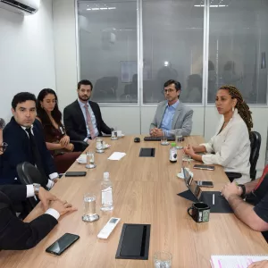 Reunião em uma sala com oito pessoas sentadas ao redor de uma mesa retangular de madeira. Elas estão em conversa atenta, com copos de água, canecas, garrafas e cadernos sobre a mesa. O ambiente é corporativo, com paredes brancas, janelas de vidro e iluminação clara. As pessoas usam roupas formais e demonstram concentração no diálogo.