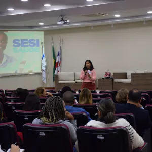 Em um auditório, uma mulher fala ao público segurando um microfone. Ela veste blusa rosa e calça marrom. À frente, há várias fileiras de cadeiras ocupadas por pessoas atentas. No fundo, há um telão com a imagem de um homem sorridente e o texto “SESI SAÚDE”. Próximo ao telão, estão três bandeiras e dois sofás de fibra com almofadas claras. O ambiente é bem iluminado e organizado, com clima de palestra ou apresentação.