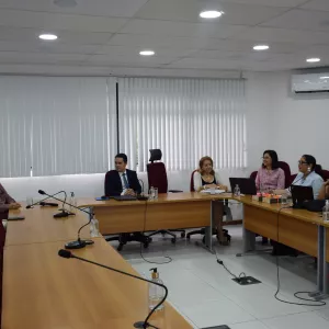 Em uma sala de reunião iluminada, oito pessoas estão sentadas ao redor de mesas em formato de “U”.  Todos parecem atentos e conversam entre si. Sobre as mesas há notebooks, garrafas de água, papéis e microfones. O ambiente tem paredes brancas, cadeiras com encosto vermelho e janelas cobertas por persianas verticais brancas.