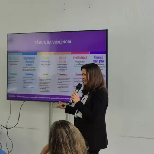 Uma mulher está em pé, segurando um microfone e apresentando em frente a uma tela grande. Ela tem cabelos castanhos, usa blazer preto e blusa clara. Na tela aparece o título “Régua da Violência”, com uma tabela colorida em cinco colunas, que classificam diferentes níveis de violência, de “Não ofensivo” até “Violência mais grave”. Algumas pessoas estão sentadas assistindo à apresentação, em um ambiente claro, com paredes brancas e janelas amplas.