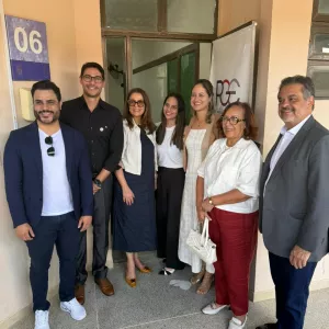 Sete pessoas estão em pé, sorrindo para a câmera, em frente a uma parede bege com uma porta de vidro e uma faixa com o logo "PGE". São quatro mulheres e três homens, vestidos com roupas sociais ou casuais elegantes. À esquerda, um homem de blazer azul e camiseta branca. Ao centro, duas mulheres usam roupas claras. À direita, uma senhora com blusa branca e calça vermelha segura uma bolsa clara, ao lado de um homem de terno cinza. Todos parecem felizes e em um ambiente profissional.