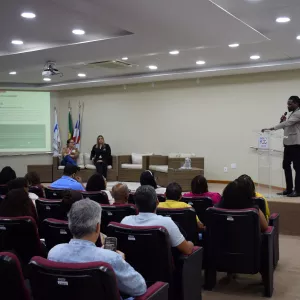 Auditório com pessoas sentadas em poltronas bordô assistindo a uma palestra. Um homem em pé, de blazer claro e calça escura, fala ao microfone em um púlpito transparente com o logotipo da PGE. No fundo, há duas mulheres sentadas em poltronas próximas a bandeiras do Brasil, da Bahia e da PGE-BA. Um telão projeta o tema “CPAD e Assessoria Jurídica na Gestão Documental”. O ambiente é bem iluminado, com teto branco e luzes embutidas.