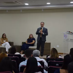 Auditório com várias pessoas sentadas em cadeiras estofadas vinho, voltadas para o palco. No palco, três pessoas estão acomodadas em sofás de fibra: um homem de terno azul, uma mulher de blazer bege e calça branca e outra de vestido azul-marinho. Em pé, à direita, um homem de terno cinza e gravata amarela fala ao microfone próximo a um púlpito transparente com o logotipo da PGE-BA. O fundo é uma parede bege lisa e o ambiente é iluminado por luzes embutidas no teto.