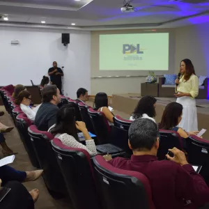 Uma mulher fala em pé diante de um auditório, segurando um microfone. Ela veste blusa amarela clara e saia branca. As pessoas estão sentadas em cadeiras vinho, observando e fazendo anotações. No fundo, há um telão com o logotipo do “Programa de Desenvolvimento de Lideranças da PGE-BA”. Um homem ao lado do auditório faz interpretação em Libras. O ambiente é iluminado e organizado, com clima de evento institucional.