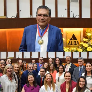 A imagem é dividida em duas partes. Na parte superior, um homem de meia-idade, com cabelo curto e óculos, veste terno azul e camisa branca. Ele sorri levemente e usa uma medalha dourada pendurada por fita vermelha e azul. Atrás dele há o símbolo da ALBA, com flores amarelas ao lado. Na parte inferior, o homem aparece novamente, ao centro, cercado por cerca de vinte pessoas, homens e mulheres, que sorriem para a foto em um ambiente interno iluminado e formal.