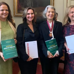 Quatro mulheres estão em pé, sorrindo, lado a lado, em um ambiente formal com quadros nas paredes. Duas delas seguram um livro verde intitulado "Estudos de Direito Público" e as outras duas seguram um livro branco com a palavra "Solidariedade" em vermelho. A terceira mulher, de cabelos grisalhos e roupa preta, usa uma medalha no pescoço. Todas estão bem-vestidas e aparentam estar em um evento jurídico-acadêmico.