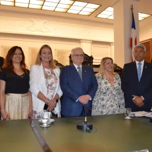Oito pessoas posam em pé atrás de uma mesa de madeira verde, com microfones e copos d’água sobre ela. Todas estão sorrindo e vestidas de forma formal, com ternos, vestidos e blazers. O ambiente é iluminado e tem bandeiras da Bahia e do Brasil ao fundo, além de detalhes clássicos nas paredes e no teto, indicando um evento institucional.