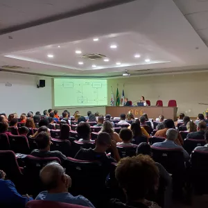 Visão geral de um auditório com uma plateia sentada em poltronas vermelhas escuras, voltada para um palco. A sala é iluminada e tem um teto rebaixado.  No palco, uma mesa escura com duas pessoas, com um pódio de acrílico à direita. Atrás delas, três bandeiras e uma tela de projeção exibem um fluxograma simples e claro. A plateia é diversificada e preenche o auditório.
