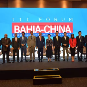 A imagem mostra quinze pessoas em um palco, sendo treze homens e duas mulheres, todas vestidas com roupas formais. Elas estão alinhadas, olhando para a câmera e sorrindo. Ao fundo, há uma grande tela azul com um banner vermelho onde se lê “III FÓRUM BAHIA-CHINA”. O ambiente é bem iluminado e o palco tem um tapete cinza escuro.