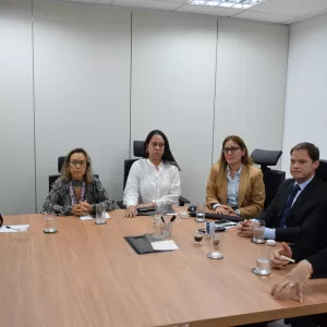 Seis pessoas estão sentadas ao redor de uma mesa de madeira em uma sala de reunião. Três mulheres estão de um lado, e uma mulher e dois homens do outro, totalizando seis pessoas. Elas olham atentamente para frente, com expressões sérias e concentradas. Sobre a mesa há copos com água, xícaras de café, celulares, cadernos e canetas. O ambiente é claro, com paredes brancas e iluminação de teto.