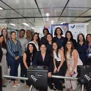 Um grupo de vinte pessoas, composto por homens e mulheres, posa sorrindo para a câmera em um ambiente interno. A maioria veste roupas sociais ou casuais escuras. O grupo está de pé, exceto por um homem no centro, agachado, e uma mulher à sua frente. Eles se agrupam próximos a duas catracas pretas, com a palavra "ENTRADA" em branco. Ao fundo, painéis e cartazes coloridos estão em exposição. As luzes de teto são fluorescentes.