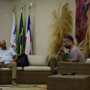 Dois homens sentados nas poltronas de fibra conversam. Um deles segura o microfone, enquanto o outro escuta com expressão atenta, com o braço apoiado na poltrona. O cenário tem bandeiras ao fundo, arranjos de folhas secas e o painel com a imagem da mulher negra. O clima é de diálogo durante um debate.