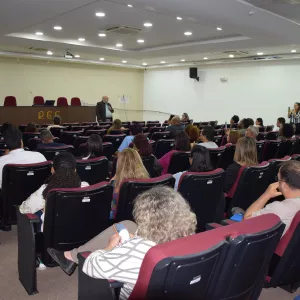 A imagem mostra um auditório com várias pessoas sentadas em poltronas vermelhas, assistindo a uma palestra. À frente, um homem de terno escuro e camisa azul fala com microfone, próximo a uma mesa com cadeiras e à parede onde estão bandeiras do Brasil, da Bahia e de outra instituição. O ambiente é bem iluminado, com teto branco e luzes embutidas. Algumas pessoas estão atentas ao palestrante, enquanto outras usam o celular ou observam em silêncio.