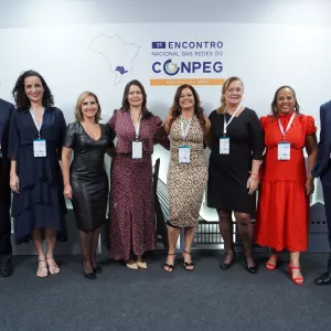  A imagem mostra oito pessoas posando para foto em frente a um painel de evento. Elas estão alinhadas lado a lado, sorrindo, e usam roupas sociais e crachás. Há homens e mulheres no grupo, todos em postura formal e amistosa. O painel atrás é claro, com logotipos repetidos. O ambiente parece ser de congresso ou encontro profissional.