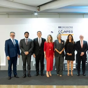 A imagem mostra um grupo de dez pessoas alinhadas lado a lado, posando para a foto em um evento. São sete homens e três mulheres, todos vestidos de forma formal, com ternos ou vestidos sociais. Eles estão em frente a um painel branco com o logotipo do “1º Encontro Nacional das Redes do CONPEG – São Paulo”. O chão é de cor neutra e o ambiente é bem iluminado. Todos sorrirem levemente, aparentando profissionalismo e cordialidade.
