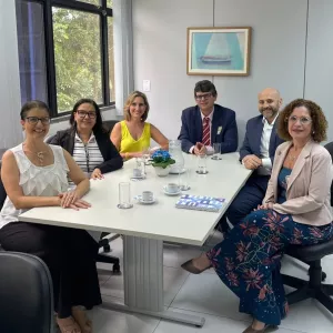 Seis pessoas estão sentadas em volta de uma mesa de reuniões branca, em um ambiente de escritório claro e organizado. Três mulheres estão de um lado e uma mulher e dois homem do outro. Todos sorriem e estão vestidos de forma social. Sobre a mesa há xícaras de café, copos de água, uma jarra, uma revista e um pequeno vaso com flores azuis. Ao fundo, há uma janela grande com vista para árvores e um quadro com a imagem de um barco à vela.