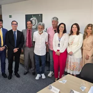 A imagem mostra um grupo de dez pessoas em pé, posando para uma foto em um ambiente de escritório. São sete homens e três mulheres. Alguns usam ternos e roupas formais, enquanto outros estão vestidos de forma mais casual. Ao fundo, há uma parede branca, um quadro, uma porta de madeira e móveis de escritório. À frente, há uma mesa com copos e papéis. Todos olham para a câmera e sorriem.