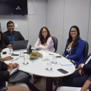 A foto mostra um grupo de pessoas sentado ao redor de uma mesa redonda branca em uma sala de reunião. Há copos de água, celulares e cadernos sobre a mesa. Todos observam alguém fora do quadro, alguns sorrindo. Ao fundo, há uma TV desligada na parede. O ambiente é claro e organizado.