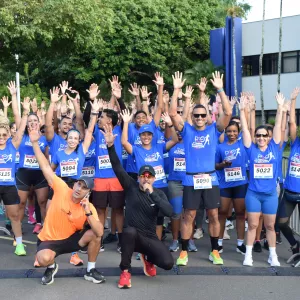 Foto que mostra um grupo de cerca de 20 corredores de rua, sorridentes, de camiseta azul com o logo da corrida. Eles estão em pé, com os braços levantados, logo após a linha de chegada de uma corrida. Dois homens estão agachados na frente.