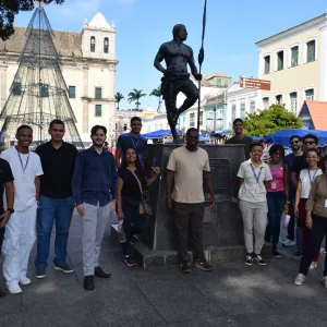 Um grupo grande posa para foto em frente à estátua de Zumbi dos Palmares, em uma praça com prédios históricos ao fundo. Todos estão alinhados ao redor do monumento, sorrindo. Há barracas de artesanato e uma estrutura de árvore de Natal próxima. O céu está azul e a luz do dia ilumina claramente o local.