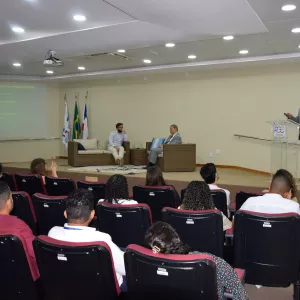 A imagem mostra um auditório onde um homem está em pé, falando em um púlpito à frente de uma plateia. Ele veste paletó cinza e segura um microfone. Ao fundo, há um telão verde com texto amarelo sobre "Principais doenças masculinas", listando várias condições de saúde. Três bandeiras estão ao lado do púlpito. Na frente do palco, dois homens estão sentados em um sofá. O público está sentado em cadeiras vermelhas e pretas, assistindo à apresentação. O ambiente é claro e formal, com iluminação no teto.