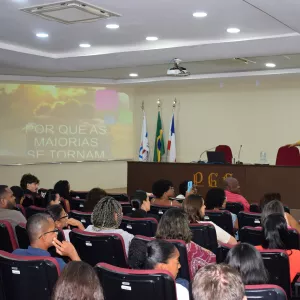 Auditório cheio, com pessoas sentadas em cadeiras acolchoadas, assistindo a uma palestra. No palco, uma mulher de roupa amarela fala ao público ao lado de uma mesa com computador e microfone. Ao fundo, um telão mostra um slide com a frase “Por que as maiorias se tornam…”. Há bandeiras posicionadas atrás da mesa e iluminação de teto ligada.