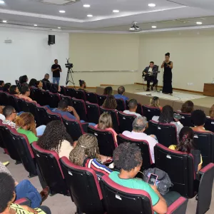 A plateia observa a apresentação no palco, onde a cantora e o músico realizam a performance. A imagem mostra a visão geral da sala, com todas as fileiras ocupadas