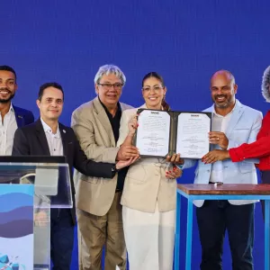 Seis pessoas, quatro homens e duas mulheres, estão em pé em frente a um painel azul. O grupo segura um documento em conjunto.  Da esquerda para a direita: um homem negro de blazer azul; um homem branco de camisa social preta; um homem branco de blazer bege; uma mulher branca de blazer e calça bege claro; um homem negro de blazer azul claro; e uma mulher negra de camisa vermelha. Todos sorriem.