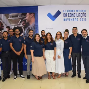 Um grupo de doze pessoas posa sorridente em um ambiente interno, diante de um grande banner azul e branco com o texto “XX Semana Nacional da Conciliação – Novembro 2025”. A maioria veste camisa polo azul-marinho com o logotipo da PGE, calças sociais ou saias em tons neutros. O grupo está lado a lado, demonstrando integração e alegria. O local tem piso claro, parede branca e cortina bege à direita.
