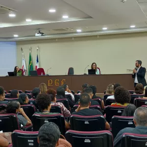 Auditório com várias pessoas sentadas em poltronas vermelhas, voltadas para uma mesa elevada onde duas mulheres estão sentadas com notebooks. À direita, um homem de terno fala em pé com microfone. Na parede ao fundo há bandeiras e um telão com apresentação projetada.