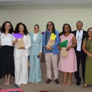 Oito pessoas posam sorridentes em frente ao palco de um auditório. O grupo está bem vestido, com roupas sociais e elegantes, e alguns seguram saquinhos de presente coloridos. Ao fundo, há bandeiras e um púlpito com o logotipo da PGE. O clima é de confraternização e encerramento de um evento.