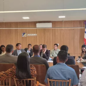 Sala de reuniões ampla e iluminada. Cerca de vinte pessoas adultas estão sentadas ao redor de uma mesa retangular, conversando e ouvindo. A maioria usa roupas sociais. Algumas pessoas anotam em cadernos ou usam notebooks e celulares. Ao fundo, há uma parede de madeira com um brasão e a palavra “Juazeiro”. À direita, três bandeiras em suportes verticais. À esquerda, quadros na parede azul. A cena mostra uma reunião formal em andamento.