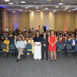 Foto geral do auditório com grande público e três pessoas em pé, ao centro, posando para a imagem. As cadeiras ocupam quase todo o espaço, mostrando alta participação. A estrutura de gravação e iluminação aparece ao fundo.
