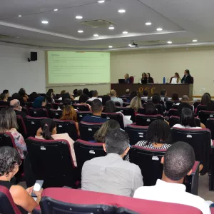 A imagem mostra um auditório cheio, com várias fileiras de cadeiras vermelhas ocupadas por pessoas adultas. Todos estão voltados para a frente, onde há uma mesa comprida com cinco pessoas sentadas, participando de uma apresentação. Um projetor exibe um slide com texto na parede. O ambiente é iluminado e organizado, com clima de palestra ou reunião formal. No canto direito, há um púlpito transparente com o logotipo “PGE”.