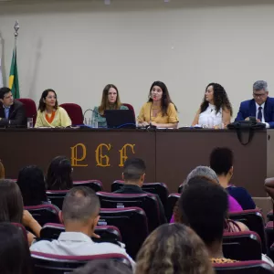 A imagem mostra seis pessoas sentadas lado a lado em uma mesa longa, participando de um evento formal. Há quatro mulheres e dois homens, todos vestidos de forma elegante. Atrás deles, estão três bandeiras: uma do Brasil, uma estadual e a terceira da PGE-BA. À frente da mesa, está a sigla "PGE" em letras grandes. Na parte inferior, algumas pessoas assistem à apresentação. O ambiente é uma sala de conferências com iluminação clara e um telão ao lado esquerdo. A cena transmite uma reunião oficial.