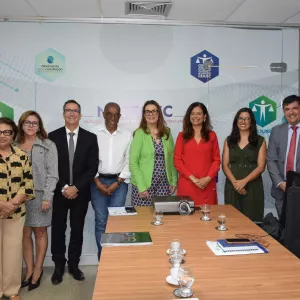 Grupo de oito pessoas em pé, dentro de uma sala de reuniões, conversando em frente a uma parede com logotipos do Poder Judiciário e do CEJUSC.
