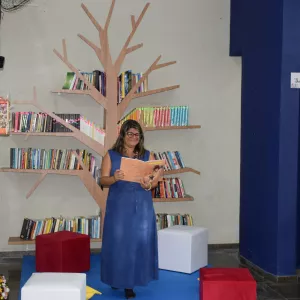 Uma mulher está em um espaço de leitura, em frente a uma estante em formato de árvore. Ela segura um livro aberto e sorri. O ambiente tem pufes coloridos, muitos livros, plantas e decoração com desenhos de pássaros. A cena transmite clima de leitura e evento cultural.