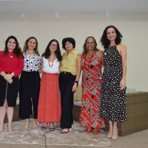 Seis mulheres posam sorrindo em frente aos sofás do auditório. Elas usam roupas coloridas e variadas, transmitindo diversidade. O ambiente é institucional e o grupo parece registrar o encerramento da atividade ou palestra