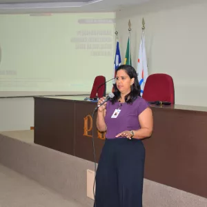 Uma mulher fala ao microfone durante uma apresentação. Ela veste blusa roxa e crachá, posicionada diante de uma tela projetada. O ambiente parece ser uma sala de reunião ou auditório institucional.