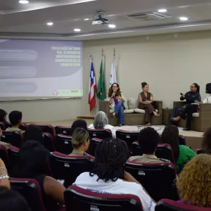 Em um auditório, várias pessoas estão sentadas em fileiras de cadeiras, formando o público e voltadas para o palco. No palco, quatro mulheres adultas estão sentadas em sofás claros, participando de uma roda de conversa. Uma delas segura o microfone e fala, enquanto as outras escutam. Ao fundo, um telão exibe informações do evento sobre fiscalização em tempo real e a Lei Maria da Penha. À direita do palco, estão as bandeiras da Procuradoria Geral, de Salvador e da Bahia. 