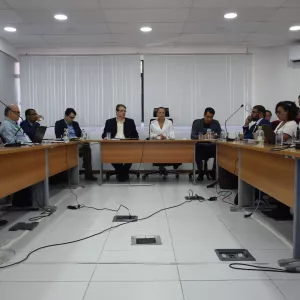 Reunião institucional em sala ampla e bem iluminada. Pessoas sentadas ao redor de mesas em formato de U, com microfones, notebooks, copos de água e documentos. Os participantes estão atentos, ouvindo e registrando informações, em ambiente formal de trabalho.