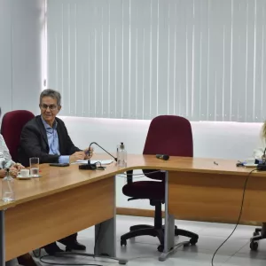 Imagem em ambiente interno, uma sala de reuniões. Três pessoas estão sentadas ao redor de uma mesa de madeira em formato angular. À esquerda, uma mulher de cabelo castanho curto usa roupa clara e conversa com um homem ao centro, que tem cabelos grisalhos, usa óculos e veste terno escuro com camisa clara. À direita, outra mulher, de cabelos longos e castanhos, veste blusa clara. Sobre a mesa há microfones, copos com água, xícaras de café e papéis. Ao fundo, parede clara com persianas verticais brancas.