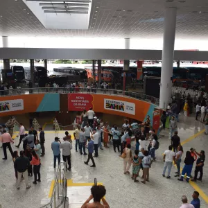 Vista ampla do interior da Nova Rodoviária da Bahia, em Salvador. O espaço é grande, com piso claro, teto alto e iluminação natural. Muitas pessoas circulam, conversam ou aguardam. Há escadas, filas organizadas e painéis com a identidade visual do Governo da Bahia. Ônibus estão visíveis ao fundo.