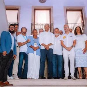Foto em ambiente interno. Em uma escadaria, há um grupo de cerca de onze pessoas adultas, lado a lado, olhando para a câmera. Elas estão em um patamar de escada, com paredes laterais decoradas com azulejos azuis e brancos. Ao fundo, há três janelas altas com grades. A maioria veste roupas claras, em tons de branco e azul. Algumas pessoas usam óculos. A iluminação é suave e o clima da imagem parece formal e coletivo.