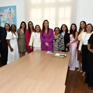 Registro de foto oficial com um grupo de mulheres e homens em uma sala de reuniões. Todos estão em pé, lado a lado, sorrindo para a câmera. O cenário é formal e representa união, representatividade e participação coletiva em um encontro institucional.