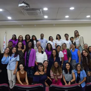 Grupo com cerca de 30 pessoas posa para foto em um auditório. A maioria é de mulheres, de diferentes idades e tons de pele, e há alguns homens. Estão organizadas em três filas, em pé e agachadas, todas sorrindo. Ao centro, uma mulher negra usa blazer roxo. Ao fundo, aparecem bandeiras e uma parede clara. No teto, há luzes embutidas e um projetor. Na frente, vêem-se cadeiras vermelhas. O ambiente é interno, iluminado e institucional.