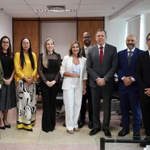 Nove pessoas estão alinhadas, posando para uma foto formal em um ambiente de escritório. São cinco mulheres e quatro homens, todos vestidos com roupas sociais. As mulheres usam blusas e calças ou saias em tons variados, incluindo amarelo, preto e branco. Os homens vestem ternos escuros com gravatas coloridas. O ambiente é claro, com paredes brancas, janelas com persianas e móveis de escritório ao fundo. Todos estão de pé, sorrindo ou com expressão séria, transmitindo profissionalismo.